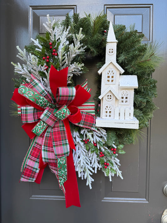 Decorative Christmas wreath with a plaid green, red, and white bow and small white church figure on a door.
