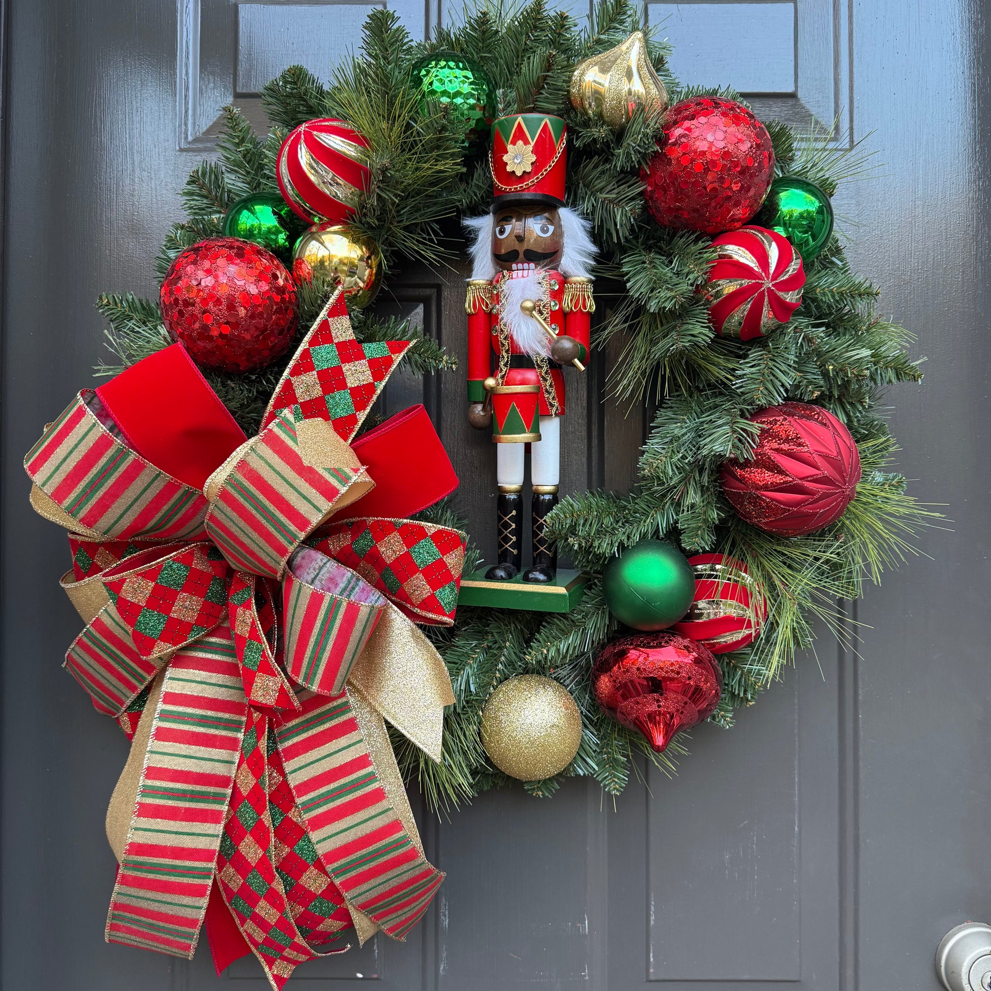 Decorative Christmas wreath with ornaments, a red, gold, and green bow, and a nutcracker figure on a door.